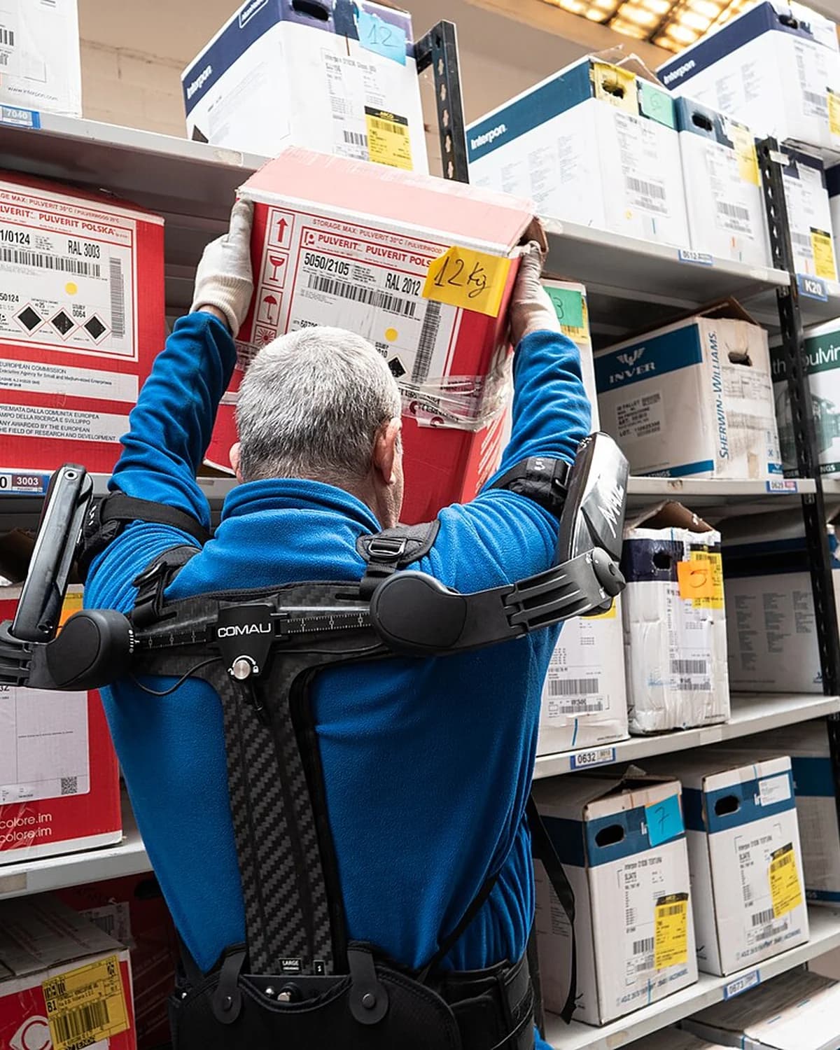 A worker lifting a box overhead while wearing the Comau MATE-XT passive exoskeleton.